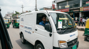 A white Tata Motors delivery van moves through a busy Bangladeshi street, its sides marked with 'GLOBAL LOGISTICS' and '24/7 TRACKED' in both English and Bengali. A smartphone is mounted on the windshield, displaying a map with a GPS location pin. The street is crowded with multiple green and yellow auto-rickshaws and other vehicles, with roadside shops featuring Bengali signboards and power lines in the background, all illustrating dynamic city life in Bangladesh.